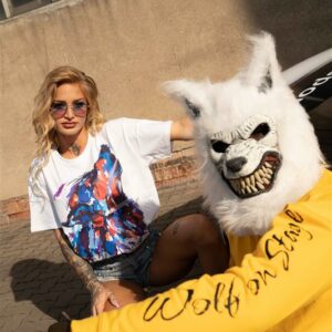 Zwei junge Erwachsene in urbaner Umgebung; im Fokus ein Mann mit weißer Wolfsmaske, der einen gelben Pullover mit der Aufschrift „Wolf on Stage“ trägt. Neben ihm sitzt eine Frau in weißem T-Shirt mit bunter Grafik – beide verkörpern Streetstyle, Rebellion und urbane Coolness.