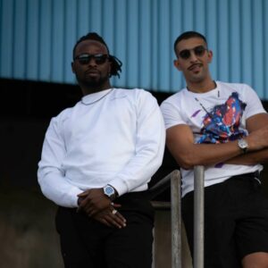 Zwei junge Männer stehen lässig nebeneinander auf einer Metallplattform vor einem urbanen Industrie-Hintergrund. Der linke trägt ein weißes Sweatshirt mit dezenter Logo-Stickerei von Wolf on Stage, schwarze Hose, schwarze Boots und Sonnenbrille. Der rechte trägt ein weißes T-Shirt mit buntem Print, schwarze Shorts, Sonnenbrille und Sneaker. Beide blicken selbstbewusst in die Kamera.