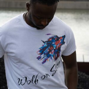 Ein Mann mit hochgesteckten, gedrehten Haaren steht draußen vor einem Gewässer und blickt nach unten. Er trägt ein weißes T-Shirt mit einem bunten, abstrakten Wolfskopf-Motiv auf der Brust. Unter dem Motiv steht in schwarzer, stilisierter Schrift: „Wolf on Stage“. Der Mann hat die Hände in den Hosentaschen und trägt eine silberne Uhr am Handgelenk sowie eine silberne Kette.